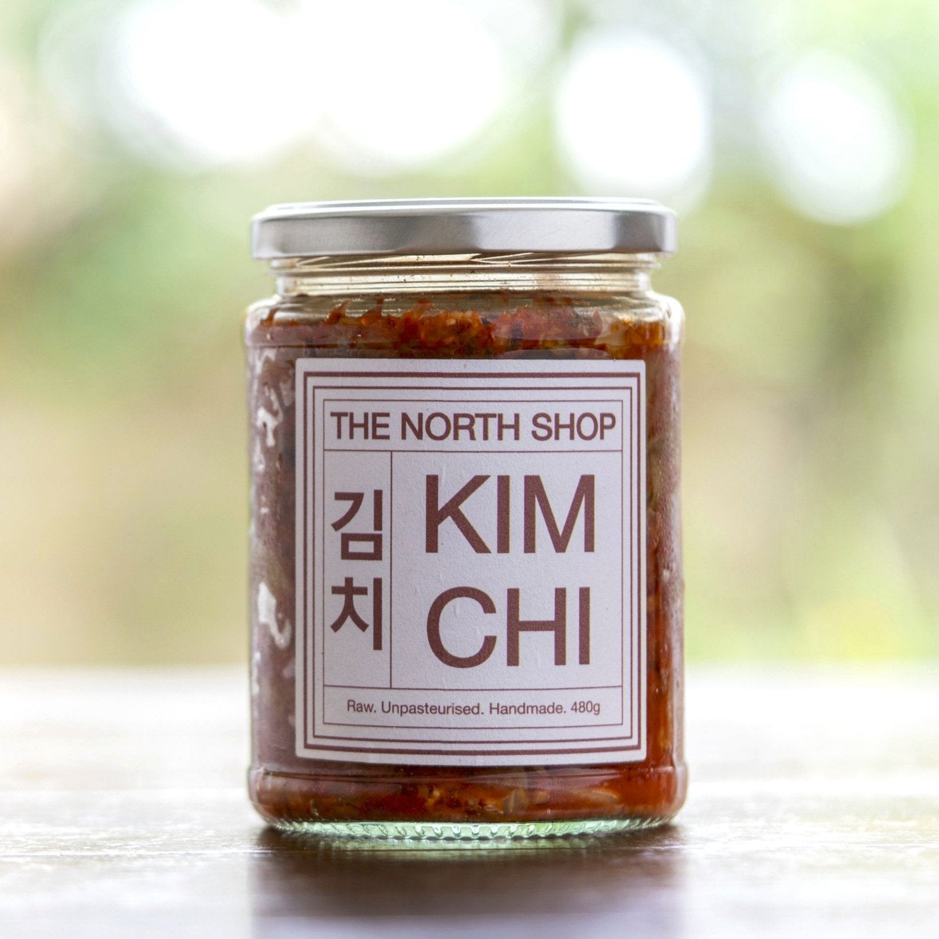 A jar of The North Shop kimchi on a wooden table with the soft image of a garden in the background.