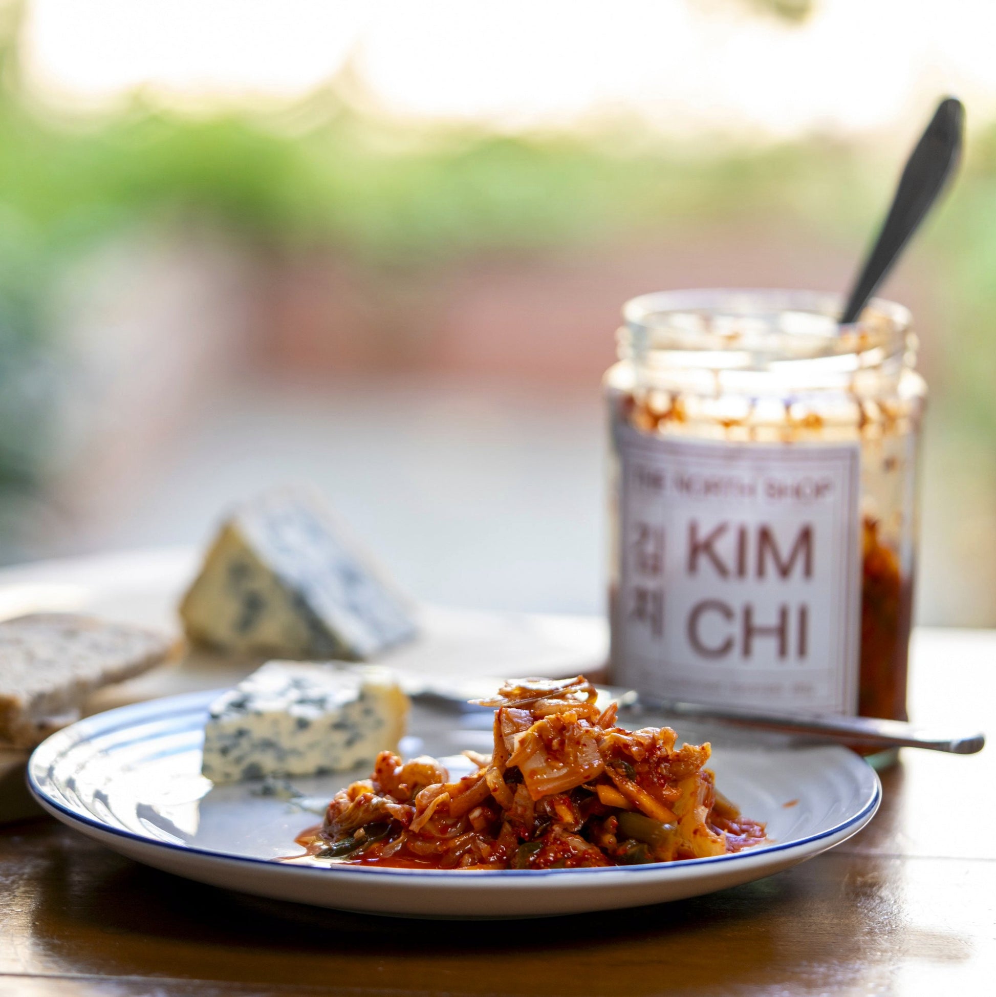 Kimchi and a piece of blue cheese on a white plate on a wooden table in front of an open jar of kimchi with a spoon in it.